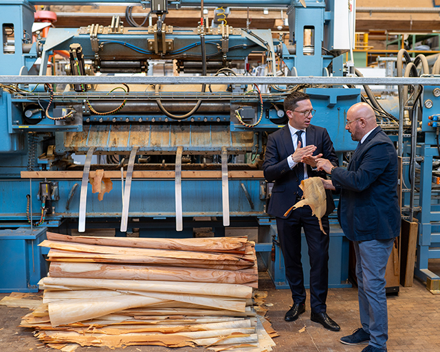 Das Foto zeigt Minister Falko Mohrs und Institutsleiter Professor Kasal vor einer großen technischen Anlage, auf der gerade Holzfurnier hergestellt wird.