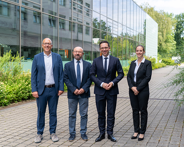 The photo shows four people on the outdoor area of the Fraunhofer Campus Braunschweig. A laboratory building with a glass front can be seen in the background.